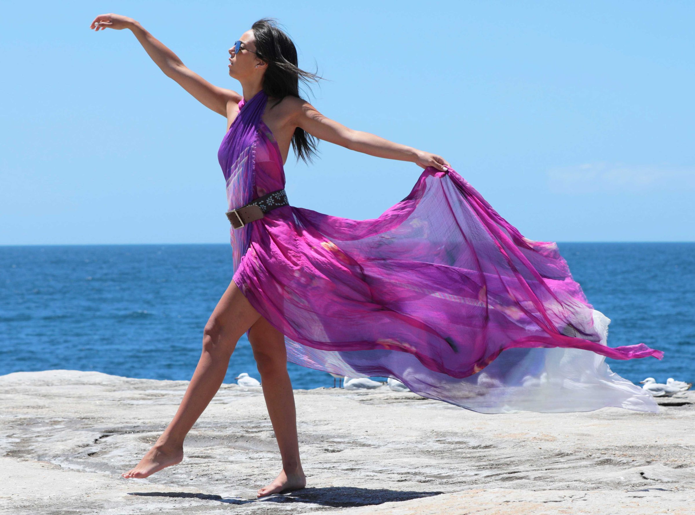 A model dancing on a rock platform above the sea in a flowing magenta nikkib sarong